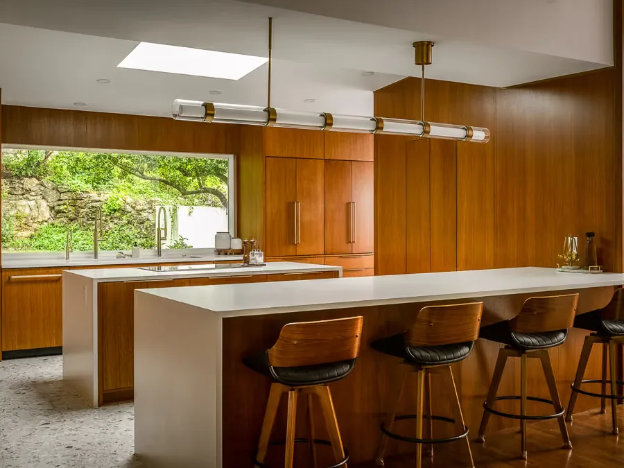 The same Balcones kitchen after work, with walnut cabinetry, pale worktops and one wide window onto the stone retaining wall.